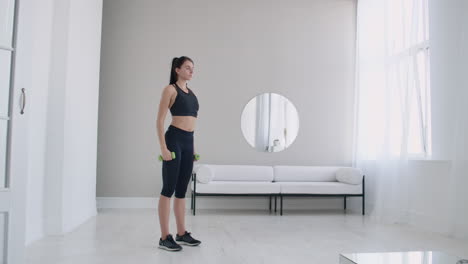 A-woman-raises-her-arms-with-dumbbells-doing-a-shoulder-exercise-in-her-light-white-apartment-against-a-sofa-and-window.